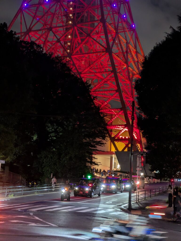 赤くライトアップされた東京タワーの脚部と、その下の交差点を走るタクシーの夜景