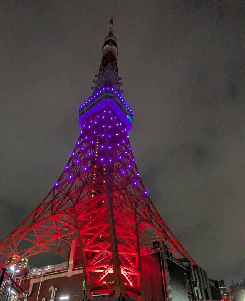 赤くライトアップされた鉄骨と紫の光の点が並ぶ東京タワーを、真下から見上げた夜景